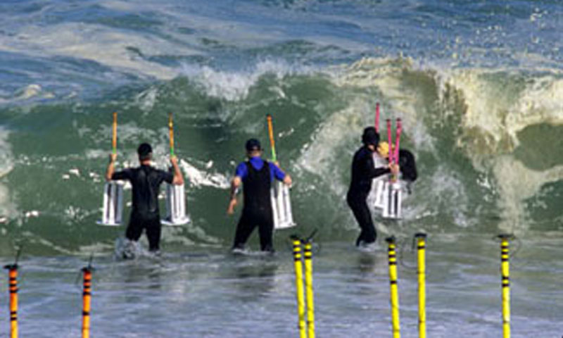 Naval Postgraduate School Oceanography Assistant Professor Jamie MacMahan and wet-suited colleagues wade into the surf zone near NPS with GPS-enabled drifters in a field experiment to track rip current flows.  Left to right:  Professor Ad Reniers of the University of Miami, MacMahan (already under the wave), Visiting Professor Nick Dodd and former students Jon Morrison and Jeff Brown.