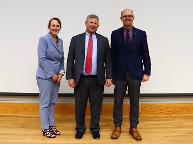 A picture of a blonde woman in grey pants suit next to grey haired man in suit with red tie next to a bald man with glasses in a blue suit in front of an auditorium